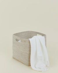 The Essential Woven Storage - Square Basket by Hawkins New York, featuring handles and a beige finish, holds a white fringed cloth draped over the side, set against a simple, light-colored background.