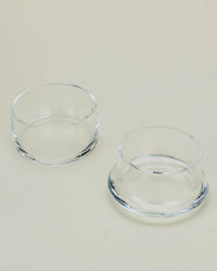 Two small, clear handblown glass bowls from Hawkins New Yorks Simple Glass Butter Keeper are placed on a light beige surface—one upright, one upside down—highlighting their smooth, minimalist design.