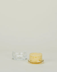A transparent Simple Glass Butter Keeper by Hawkins New York sits beside a small round yellow marbled candle on a matching glass base, both displayed against a light, neutral background.