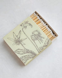 A partially opened box of Hudson Valley Matches | Trout Lillies by The Floral Society rests on a light surface, displaying illustrated matchboxes with white tips and a sketch-style botanical design of two blooming flowers and long leaves.