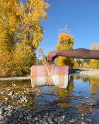 A hand holds the caminito Suave Miel - Blanket Roll, a handwoven pastel blanket with leather straps, over a rocky riverbed surrounded by golden autumn trees beneath a clear blue sky.