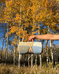 A hand holds the Suave Campo - Blanket Roll by caminito, featuring a rolled-up, multicolored design with a tan leather strap, set against tall autumn trees under a clear blue sky.