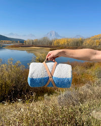 A hand holds the caminito Suave Aire - Blanket Roll, wrapped in a leather strap, with a lake, autumn trees, grass, and distant mountains under a clear blue sky in the background.