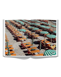 The open book "Italian Summer" by Simon & Schuster shows rows of orange lounge chairs and green-and-white umbrellas on stone, viewed from above—capturing the charm of Italy’s coastal towns.