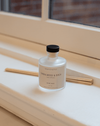 A Near & Native Room Diffuser in Sandlewood & Birch, featuring a frosted glass bottle with black cap, sits on a white windowsill next to reed sticks and a beige card. A window and blurred brick wall appear in the background.