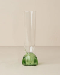 The Saarde Kairos Champagne Flute in Green, featuring a tall, clear design with a textured green base, is set upright on a light-colored surface against a plain background.
