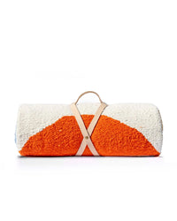 The caminito Luna Paseo - Blanket Roll is a lightweight, textured outdoor blanket in orange and white, rolled up and secured with a handmade leather strap and handle, shown against a plain white background.