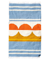 The Luna Paseo - Blanket Roll by caminito is a rectangular woven rug with blue and white stripes, orange semicircles in the center, white and yellow bands, fringed edges, is lightweight, machine washable, and perfect for outdoor use.