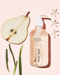 A clear pump bottle of Rosie Jane Leila Lou Everyday Body Wash sits beside a sliced pear, three white flowers, and green plant stems on a pale pink background.