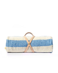 The Marea Paseo - Blanket Roll by caminito is a blue and white striped, handwoven Mexican picnic blanket, rolled up and secured with a tan leather strap and handle, shown against a white background.