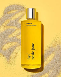 A clear glass bottle of Madie Everyday Hydrating Body Oil by Rosie Jane, featuring a silver cap and yellow formula, sits on a sand-dusted yellow surface.