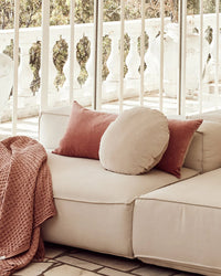 A beige sectional sofa with two pink rectangular cushions, a Saarde Linen/Velvet Round Pillow with Feather Insert in Clay, and a textured pink throw, set on a patio with white railings.