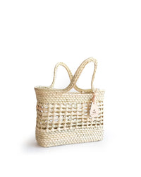 The Canasta Mini Bag in Natural by caminito is a handmade woven straw tote with two handles and a small tan leather tag, shown against a white background.