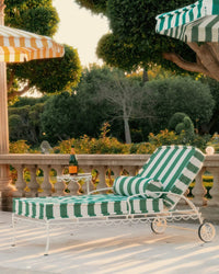 A Bolster Pillow - Crew Canyon Green Stripe by Business & Pleasure Co sits on a sunny patio lounge chair beside a small table with a bottle and glass, surrounded by lush trees and a stone balustrade.