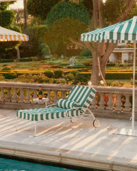 A Bolster Pillow - Crew Canyon Green Stripe by Business & Pleasure Co rests on a striped lounge chair by the pool, shaded by a matching umbrella. A side table holds two glasses and champagne amid manicured gardens, creating a relaxing scene.