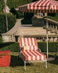 A Bolster Pillow - Crew Le Sirenuse Stripe by Business & Pleasure Co rests on a red and white striped lounge chair beneath a matching umbrella on grass, with a folded towel and drink nearby. Ornate stone stairs and manicured hedges are in the background.