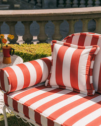 A white outdoor bench features Business & Pleasure Cos Small Square Throw Pillow in Crew Le Sirenuse Stripe, paired with matching cushions beside a round table with a red vase of yellow flowers. Stone railing and trimmed hedges complete the scene.