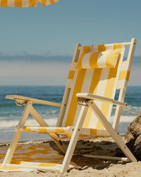 The Business & Pleasure Co Tommy Chair in Acacia White with Crew Mimosa Yellow Stripe rests on the sand by the ocean, paired with a matching towel and umbrella. Its UV resistant canvas stays vibrant against the backdrop of sea and sky.