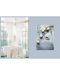 A table set by large windows with sheer curtains displays a tiered white cake from *Queen of the Cakes – Gluten-Free Baking and Artistry* by Cozy Publishing, alongside lemon layer cakes on a blue-gray surface, perfect for exploring new decorating techniques.