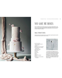 A two-tier white cake topped with pink fondant roses sits on a stand beside the book "Queen of the Cakes – Gluten-Free Baking and Artistry" by Cozy Publishing, featuring recipes and tips for crafting delicate fondant roses.