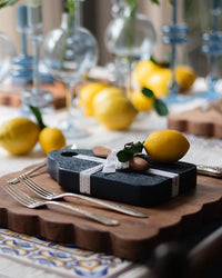 A table setting features the etúHOME Dutch Small Cheese Board w/ Spreader in Navy Blue, tied with ribbon and topped with a lemon, surrounded by cutlery, lemons, and decorative glassware.