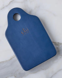 A Dutch Small Cheese Board w/ Spreader in Navy Blue by etúHOME, featuring a rounded handle with a hole, rests on a white marble surface.