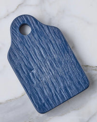 The etúHOME Dutch Small Cheese Board w/ Spreader in Navy Blue features a rectangular wood grain design with a corner hole, displayed on a white marble surface.