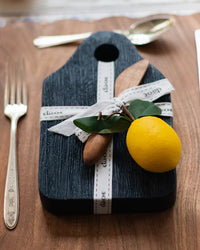 The etúHOME Dutch Small Cheese Board w/ Spreader in Navy Blue, tied with a white ribbon and topped with wooden utensils, green leaves, and a yellow lemon, sits next to a fork and knife on a wooden surface.