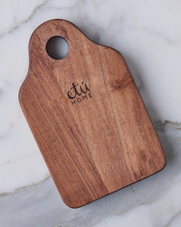 The Dutch Small Cheese Board w/ Spreader in Natural by etúHOME, featuring a rounded handle with a hole and engraved branding, rests on a light marble surface.