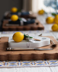 A lemon, greenery, and a wrapped utensil sit on the Dutch Small Cheese Board w/ Spreader in White by etúHOME, atop a wooden tray with more similar setups in the background.