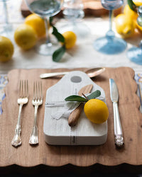 A table setting featuring etúHOMEs Dutch Small Cheese Board w/ Spreader in White, silverware, a lemon with leaves, a wooden-handled knife, and glasses, all arranged on a wooden placemat.