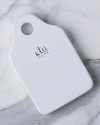 The etúHOME Dutch Small Cheese Board w/ Spreader in White is a small, rectangular ceramic serving board with rounded corners, a handle hole, and etú HOME printed on it, styled after classic Dutch cheese boards.