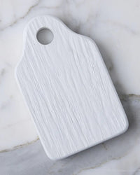 The etúHOME Dutch Small Cheese Board w/ Spreader in White features a rectangular wooden design with a handle and round hole, displayed on a light marble surface.