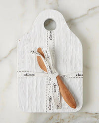 The etúHOME Dutch Small Cheese Board w/ Spreader in White features a textured board with a top hole, paired with a wooden spoon and spreader tied by a white ribbon, all displayed on marble.