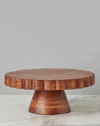 The etúHOME Natural Scalloped Cake Stand, featuring a wide, sturdy base and scalloped edge in acacia wood, sits on a marble surface against a plain gray background.