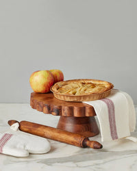 A baked apple pie rests on the etúHOME Natural Scalloped Cake Stand, surrounded by two apples, a striped kitchen towel, a rolling pin, and an oven mitt atop a marble countertop.