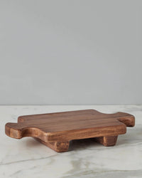 The etúHOME Normandy Wood Riser Footed Tray, Small, is a rectangular Acacia wood tray with short legs and wide handles at each end, shown on a marble surface with a gray background. Ideal as a serving board or countertop riser.