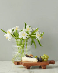 A clear glass vase with white flowers sits beside the etúHOME Normandy Wood Riser Footed Tray, Small, which holds cheese, figs, and green grapes on a white surface against a gray wall.