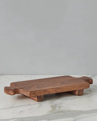 The Normandy Wood Riser Footed Tray, Medium by etúHOME features short legs and end handles, crafted from Acacia wood and elegantly displayed on a white marble surface with a gray backdrop.