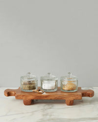 Three glass jars with lids, filled with assorted spices, are displayed on the medium Normandy Wood Riser Footed Tray by etúHOME, placed on marble against a plain gray background.