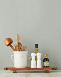 An etúHOME Normandy Wood Riser Footed Tray holds a ceramic crock with wooden utensils, two white grinders, olive oil, and vinegar on a marble kitchen countertop.