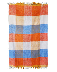 The Suave Paseo - Blanket Roll by caminito is a colorful, rectangular handwoven throw with orange, blue, white, and yellow checkered squares and short fringes on both ends—ideal for indoors or outdoors.