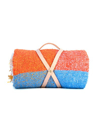 The Suave Paseo - Blanket Roll by caminito, featuring orange and blue handwoven sections, is rolled up and secured with a beige leather carrying strap and handle against a white background.