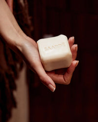 A hand holds a rectangular Saarde Olive Oil Bar Soap in Medina, embossed with the brand name, against a dark background.