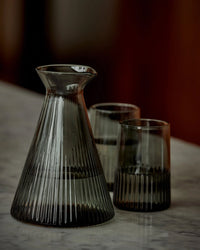 The Saarde Kairos Angular Carafe in Smoke, crafted from durable borosilicate glass with striking vertical ribbed patterns, includes two matching glasses and is displayed on a marble surface.