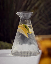 The Saarde Kairos Angular Carafe in Clear, filled with water, lemon slices, and mint leaves, rests on a white surface against a softly blurred background.