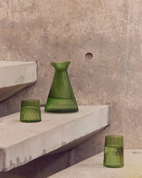The Saarde Kairos Angular Carafe in Green with two matching green glasses sits on concrete steps against a textured concrete wall.