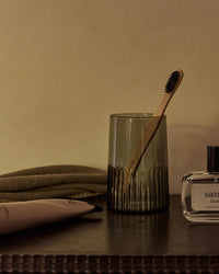A wooden toothbrush in a Saarde Kairos Large Water Glass in Smoke, with a folded towel, tube, and bottle from the same collection, are arranged on a dark countertop against a beige wall.