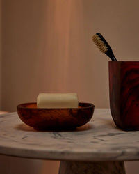 A Saarde Olive Oil Bar Soap in Andalusia rests in a brown dish, with a toothbrush in a matching cup, displayed on a round marble surface against a beige background.