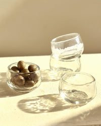 The Collective Pebbled Condiment Bowls – Set of 4, shown on a sunlit surface with three empty bowls and one filled with green olives, casting soft shadows.